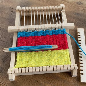 May include: A rectangular wooden loom frame with vertical white warp threads. The woven fabric features horizontal bands of bright blue, red, and yellow yarn. A wooden shuttle wrapped with blue yarn rests on the fabric, and a separate wooden comb lies nearby.