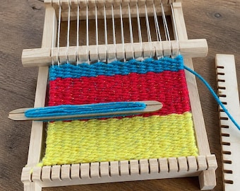 May include: A rectangular wooden loom frame with vertical white warp threads. The woven fabric features horizontal bands of bright blue, red, and yellow yarn. A wooden shuttle wrapped with blue yarn rests on the fabric, and a separate wooden comb lies nearby.