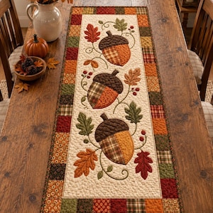 May include: A rectangular quilted table runner featuring a central cream-colored panel with three large appliqued acorns and autumn leaves in shades of orange, brown, and green, surrounded by a border of small, multi-colored patchwork fabric squares.