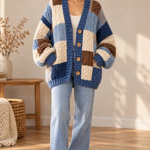 May include: A chunky knit cardigan featuring a patchwork design of blue, cream, and brown squares with varying textures including cable knit and bobble patterns. The cardigan has a blue ribbed V-neck collar, cuffs, and hem, with four large wooden buttons.