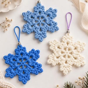 May include: Three crocheted snowflake ornaments featuring intricate yarn patterns. One snowflake is light blue with a matching loop, one is a vibrant royal blue with a blue loop, and one is off-white with a purple hanging loop and silver hardware.