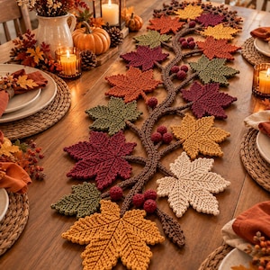 May include: A crochet table runner featuring a central brown vine-like branch adorned with various maple leaves in shades of orange, yellow, red, green, and cream, accented with small clusters of dark red berries throughout the design.