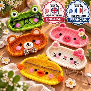 May include: Five oval crochet trays shaped like animals including a green frog, orange bear with a bow tie, pink pig with a flower, white cat with a bow, and yellow chick. Each tray features embroidered facial details and holds eyeglasses.