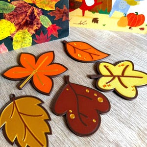 May include: Five distinct leaf-shaped ornaments in autumn colours including orange, yellow, gold, and dark brown. Each piece features embossed vein details and a smooth finish, arranged on a surface near a book with fall-themed illustrations.