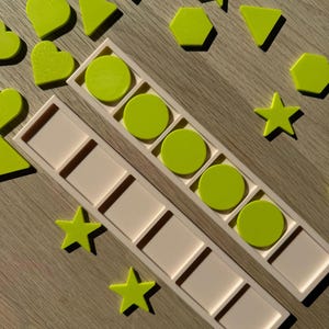 Peut inclure: Deux plateaux rectangulaires beiges avec six compartiments carrés chacun, accompagnés de diverses formes en plastique vert citron, notamment des cercles, des étoiles, des cœurs, des triangles et des hexagones dispersés sur une surface en bois clair.