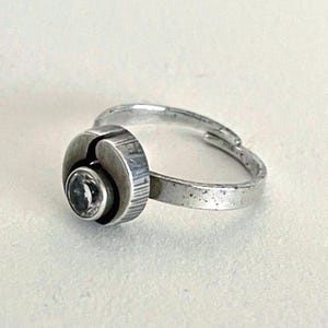 May include: A silver-toned adjustable ring featuring a circular setting with a textured, brushed finish. The center of the ring holds a single, clear, round-cut gemstone. The band is open at the back, allowing for size adjustment to fit various fingers.