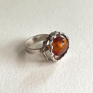 Può includere: Un anello in argento con una fascia liscia e lucida che si trasforma in una montatura testurizzata dalla forma organica. Questa montatura racchiude una singola pietra d'ambra traslucida, arrotondata e di colore arancione intenso.