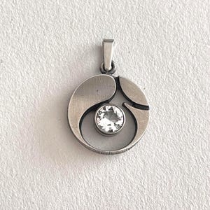 May include: A circular silver-toned pendant featuring a brushed metal finish with a central, round, clear faceted gemstone set in a bezel. The pendant has a stylized, open-work design with curved segments and includes a small, rectangular bail at the top.