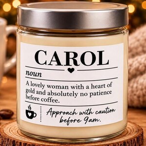 Puede incluir: Una vela en frasco de vidrio transparente con tapa plateada y cera color crema. La etiqueta blanca presenta el texto CAROL, noun, A lovely woman with a heart of gold and absolutely no patience before coffee, y Approach with caution before 9am.
