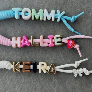May include: Three elastic bracelets featuring letter beads. The top bracelet is light blue with the name TOMMY. The middle bracelet is pink with the name HALLIE and a strawberry charm. The bottom bracelet is white with the name KEIRA and a bow charm.