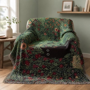 Puede incluir: Una manta tejida sobre un sillón, con un fondo verde oscuro, un patrón de flores rojas y naranjas y la silueta prominente de un gato negro con dos grandes ojos blancos que sobresalen del diseño floral.