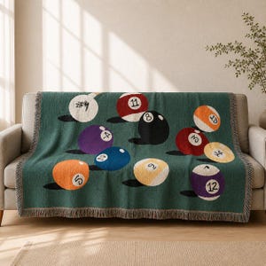 May include: A dark green woven throw blanket featuring a repeating pattern of colourful billiard balls, each marked with a unique number. The design includes balls in shades of orange, purple, blue, yellow, red, black, and white with fringed edges.