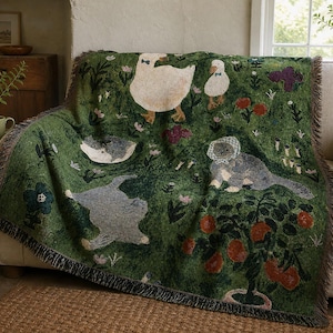 Puede incluir: Una manta tejida con fondo verde oscuro y un patrón de gansos blancos con pajaritas azules, gatos grises y un gato con collar de flores. El diseño incluye árboles frutales de color naranja, pequeñas flores y un borde con flecos.