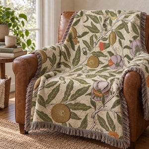 May include: A woven throw blanket featuring a botanical pattern of green leaves, brown branches, and stylized fruits in shades of yellow, orange, and purple on a cream background, finished with a decorative fringe along the edges.