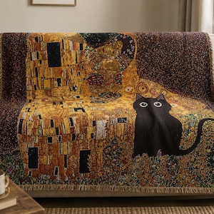 Puede incluir: Una manta tejida que presenta una interpretación estilizada de El Beso de Gustav Klimt, incorporando un gran gato negro de ojos grandes en el diseño dorado, marrón y estampado. El tejido rectangular tiene un acabado de borde con flecos.