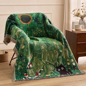 Op de afbeelding: Een geweven plaid met een levendig groen botanisch patroon, een grote zonnebloem aan de bovenkant en een kleine zwarte kat met grote witte ogen aan de onderkant. De deken heeft een textuur en decoratieve franjes langs de randen.