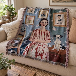 Op de afbeelding: Een geweven plaid met een centraal portret van een persoon in een rood-wit gestreept shirt, vergezeld door een zwart-witte hond. De blauwe achtergrond toont ingelijste kunstwerken en een decoratieve rand met gerafelde zijkanten.