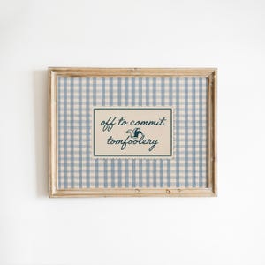 May include: A rectangular wooden frame containing a print with a light blue and white gingham pattern. Centered is a cream-coloured scalloped rectangle featuring the dark blue text "off to commit tomfoolery" with a small jester hat icon.