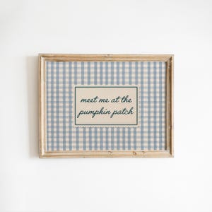 May include: A rectangular wooden frame containing a print with a light blue and white gingham pattern. Centered on the print is a cream-colored rectangular label with a scalloped edge, featuring the dark green text: meet me at the pumpkin patch.