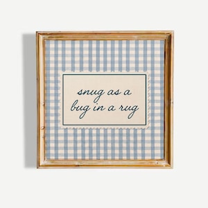 May include: A square wall art piece in a light brown wooden frame featuring a blue and white gingham pattern background. Centered is a cream-colored rectangular plaque with scalloped edges and a dark blue border, displaying the text "snug as a bug in a rug" in a dark blue cursive script.