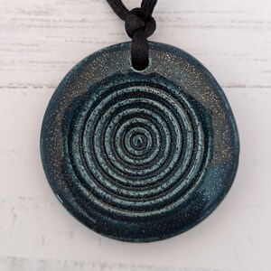 May include: A teal ceramic pendant with a spiral design and a black cord. The pendant has a textured surface with a speckled, metallic finish around the edges. The spiral pattern is centered and raised.