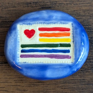 May include: A blue ceramic stone with a white rectangle painted on it. The rectangle has a rainbow flag design with a red heart in the top left corner.