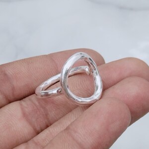 Könnte beinhalten: Ein silberner Ring mit einem unregelmäßigen, organischen Design in Form einer offenen Schlaufe und einer polierten, reflektierenden Oberfläche. Die Ringschiene weist eine leicht unebene, handgefertigte Textur auf, die eine asymmetrische Form bildet.
