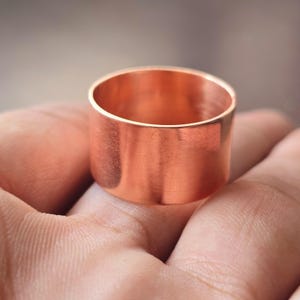 Pode incluir: Um anel largo de cobre polido com uma superfície lisa e plana e um tom castanho-avermelhado metálico consistente. A banda apresenta um design cilíndrico simples e sem adornos com bordas retas e limpas, exibindo o brilho natural do metal.