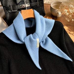 Puede incluir: Una bufanda de punto azul claro con forma triangular y una pequeña etiqueta rectangular con el texto 100%WOOL. La bufanda está colocada sobre un suéter negro, mostrando un tejido suave y texturizado con extremos puntiagudos que se superponen.