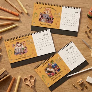 Puede incluir: Tres calendarios de escritorio con espiral que presentan ilustraciones de gatos y texto. Julio muestra un gato con una computadora portátil y el texto I NEED A SIX-MONTH VACATION TWICE A YEAR. Agosto muestra un gato con gafas y el texto I'M NOT UNDERPERFORMING. I'M SETTING REALISTIC EXPECTATIONS. Septiembre muestra un gato con el texto LABOR DAY CELEBRATING LABOR BY AVOIDING IT.
