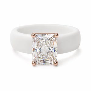 Op de afbeelding: Een ring met een gladde, matwitte band en een grote, rechthoekige, heldere steen in radiant-slijpvorm, gezet in een vierpotige zetting in roségoudkleur. De centrale steen heeft meerdere facetten die het licht reflecteren op de witte band.