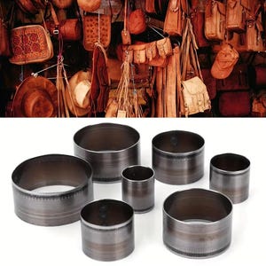 May include: A collection of seven cylindrical metal cutting dies of varying diameters, each featuring a smooth dark grey finish with a reinforced top edge and a sharp, serrated bottom cutting rim, arranged in a cluster on a white surface.