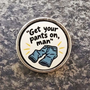 May include: A circular pin featuring a white background with the black text "Get your pants on, man" above a graphic illustration of blue jeans. The design is accented by small yellow lines radiating from the jeans, all encased in a silver metal frame.
