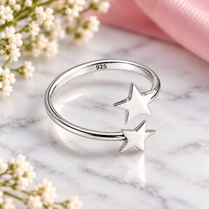 May include: A silver open-style ring featuring two polished star-shaped charms at the ends of the band. The inner surface of the band is clearly stamped with the number 925, indicating the silver content of this piece of jewelry.