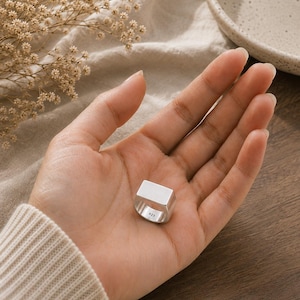 Puede incluir: Un anillo de sello de plata con una superficie superior plana y rectangular. El interior de la banda está claramente marcado con el sello 925, lo que indica una composición de plata esterlina. El anillo presenta un acabado liso y pulido.