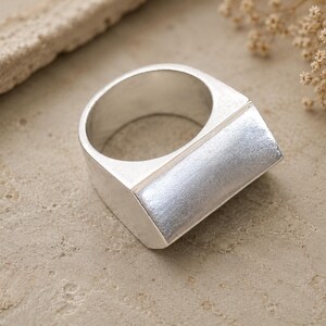 Puede incluir: Un anillo de plata que presenta una superficie superior ancha, plana y rectangular con un acabado cepillado y una banda lisa y pulida. El anillo tiene un diseño geométrico y arquitectónico con líneas limpias y una apariencia sólida y sustancial.