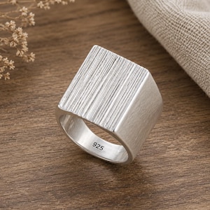 May include: A silver ring featuring a wide, rectangular top surface with a vertical ribbed texture. The band has a smooth, brushed finish and the interior is clearly stamped with the number 925, indicating the sterling silver composition of the piece.