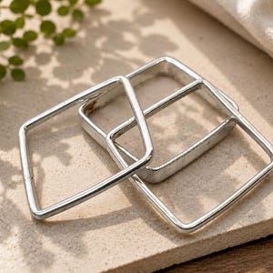 Könnte beinhalten: Drei silberfarbene, quadratische Metallrahmen mit abgerundeten Ecken. Die Rahmen sind hohl und haben eine glatte, reflektierende Oberfläche. Sie sind in einem überlappenden, leicht versetzten Muster angeordnet und bilden eine geometrische Komposition.