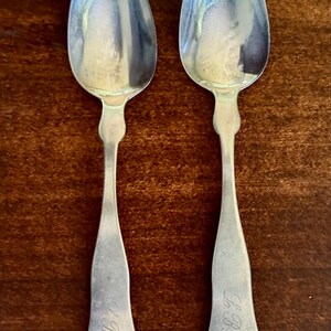 May include: Two antique silver spoons with oval bowls and long, slender handles. Each spoon has an engraved monogram, "L.B.", on the handle. The spoons are displayed on a dark wooden surface.