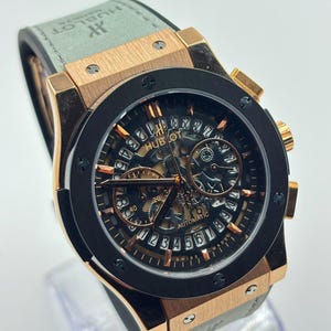 May include: A Hublot Geneve automatic wristwatch featuring a rose gold case, a black circular bezel with six screws, and a skeletonised dial with a date ring displaying numbers 1 through 31. The watch has a light grey strap with the Hublot logo.