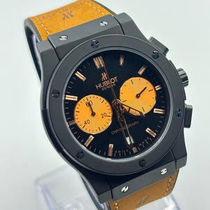 Op de afbeelding: Een Hublot Geneve chronograaf horloge met een matzwarte kast en bezel voorzien van zes H-vormige schroeven. De zwarte wijzerplaat bevat twee oranje sub-wijzerplaten, oranje uurmarkeringen en wijzers. Het heeft een beige suède horlogeband.