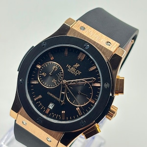 May include: A Hublot Geneve chronograph watch with a black dial, rose gold-tone case, and a black rubber strap. The watch face features rose gold-tone hour markers, hands, two sub-dials, a date window at the six o'clock position, and the text Hublot Geneve.