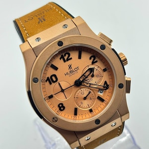 Op de afbeelding: Een Hublot Geneve chronograaf horloge met een roségoudkleurige kast en een bijpassende roségoudkleurige wijzerplaat. Het horloge heeft zwarte Arabische cijfers, drie sub-wijzerplaten, een datumvenster op de vier-uur-positie en een beige, gestructureerde lederen band.
