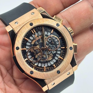Op de afbeelding: Een automatisch Hublot-horloge met een geborstelde roségouden ronde kast met zes zichtbare schroeven op de lunette. Het horloge heeft een zwarte opengewerkte wijzerplaat met een datumring, goudkleurige wijzers en een zwarte rubberen horlogeband.