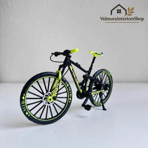 May include: A miniature mountain bike model featuring a black frame with bright neon yellow accents on the handlebars, front fork, seat, and tyre rims. The black tyres are marked with the text MTB in white, and the frame displays the word SHIMANO.