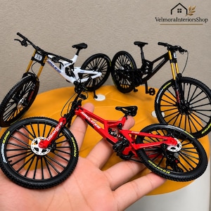 Op de afbeelding: Drie miniatuur mountainbike modellen in rood, wit en zwart, elk met gedetailleerde frames, dunne zwarte spaakwielen met gele belettering, goudkleurige voorvorken en sturen met kleine nummerplaatjes bevestigd.