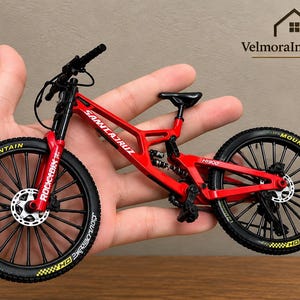 Può includere: Un modellino di mountain bike rosso con pneumatici neri e scritte gialle, sella nera e manubrio nero. Il telaio presenta la scritta bianca Santa Cruz e H900, mentre la forcella anteriore riporta il marchio bianco Rockshox.