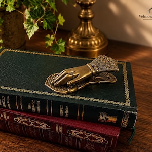 May include: A brass-colored metal hand-shaped clip with intricate filigree detailing on the cuff and a small ring on the finger, resting on top of a dark green hardcover book which is stacked on a burgundy book with gold-embossed spine lettering.