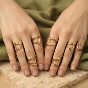 May include: A set of gold-toned rings worn on multiple fingers. Each ring features a smooth, polished metal band that wraps around the finger in an open, geometric design with a distinct triangular or angular loop shape on the outer surface.