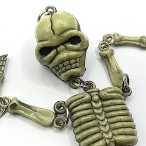 May include: A detailed, articulated skeleton keychain in a light green color. The skeleton's head, rib cage, arms, and legs are connected with metal rings. The keychain is a novelty item.